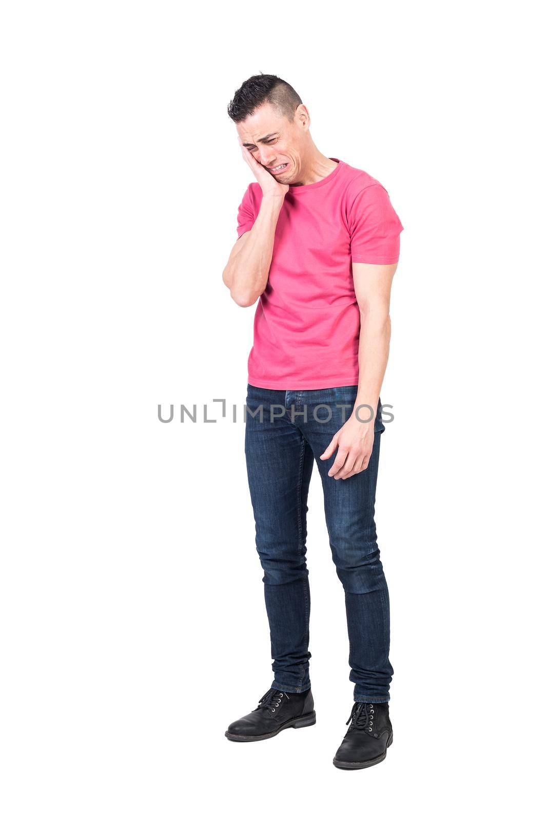 Full body of sorrowful young male with dark hair in casual clothes touching cheek and crying against white background