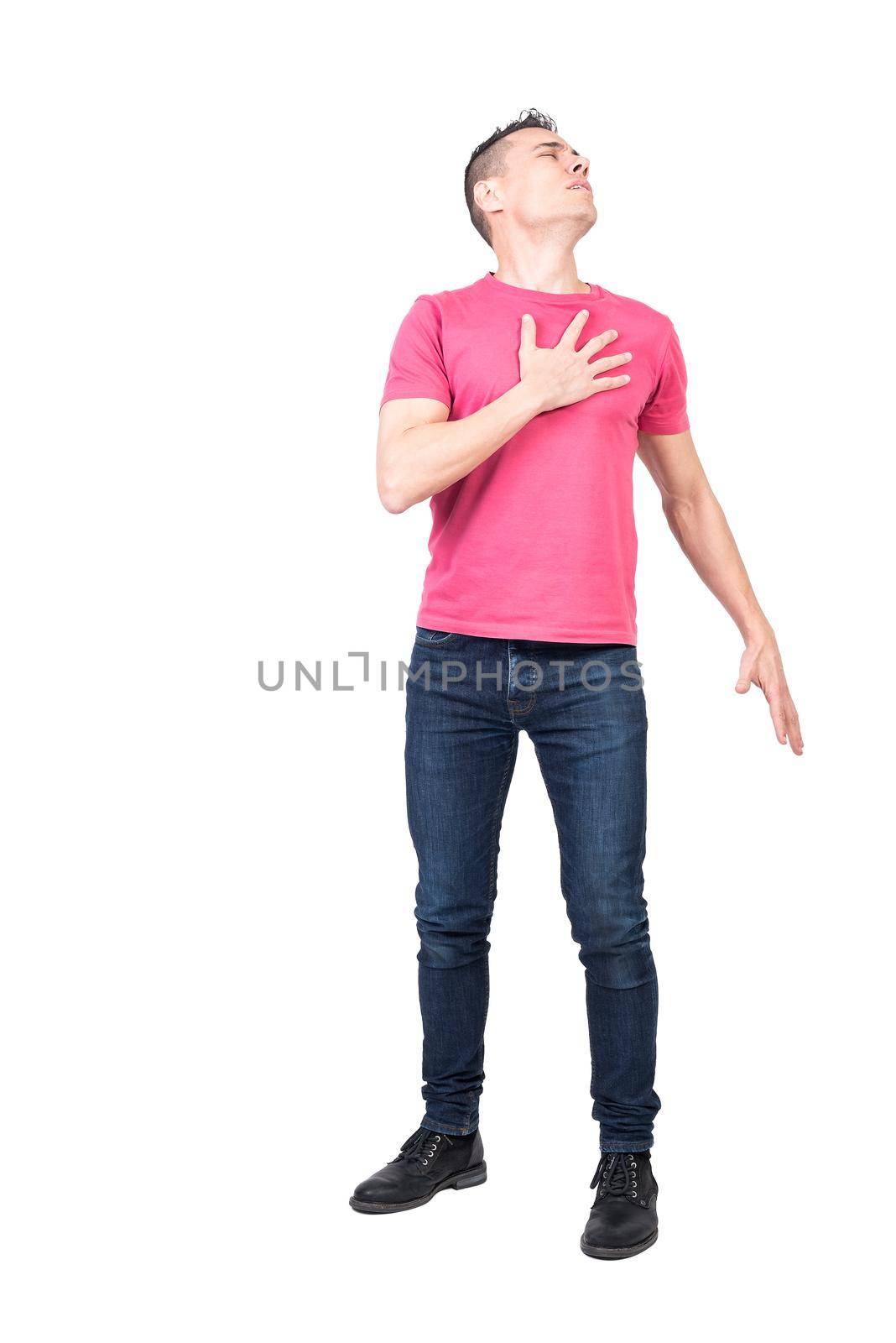 Full body of upset young male in casual clothes touching heart with closed eyes while standing against white background