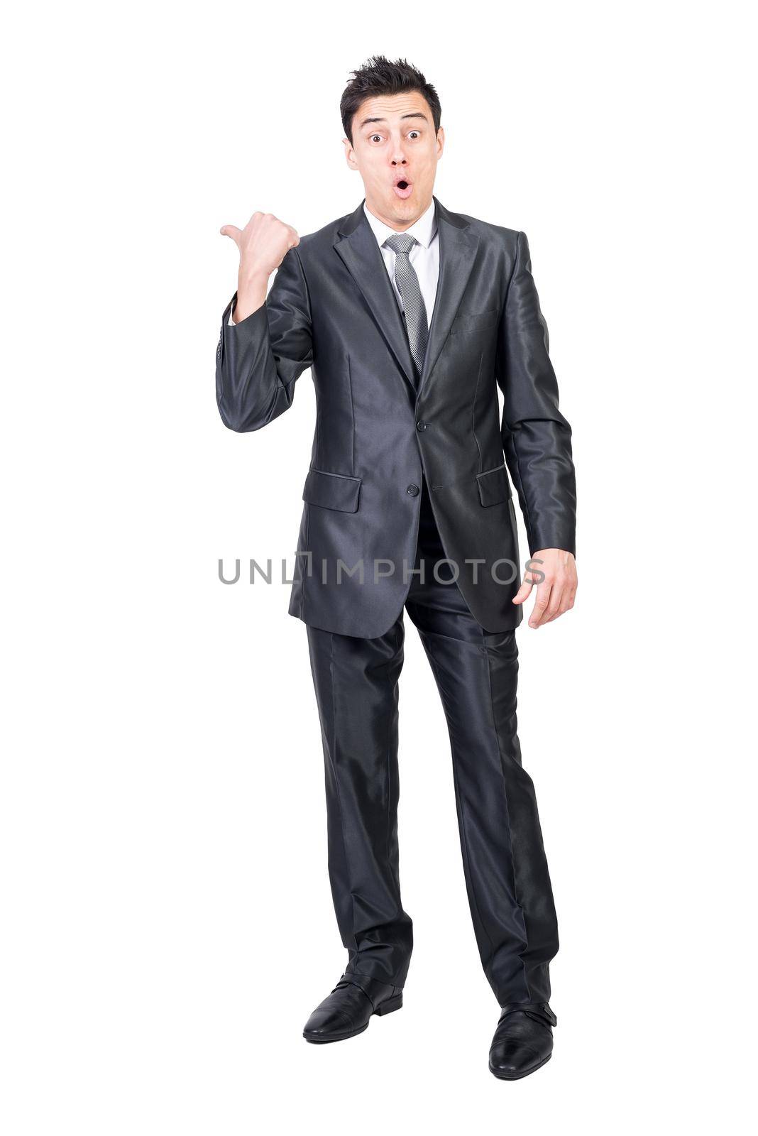 Full body of astonished male in elegant suit pointing aside with thumb and looking at camera isolated on white background