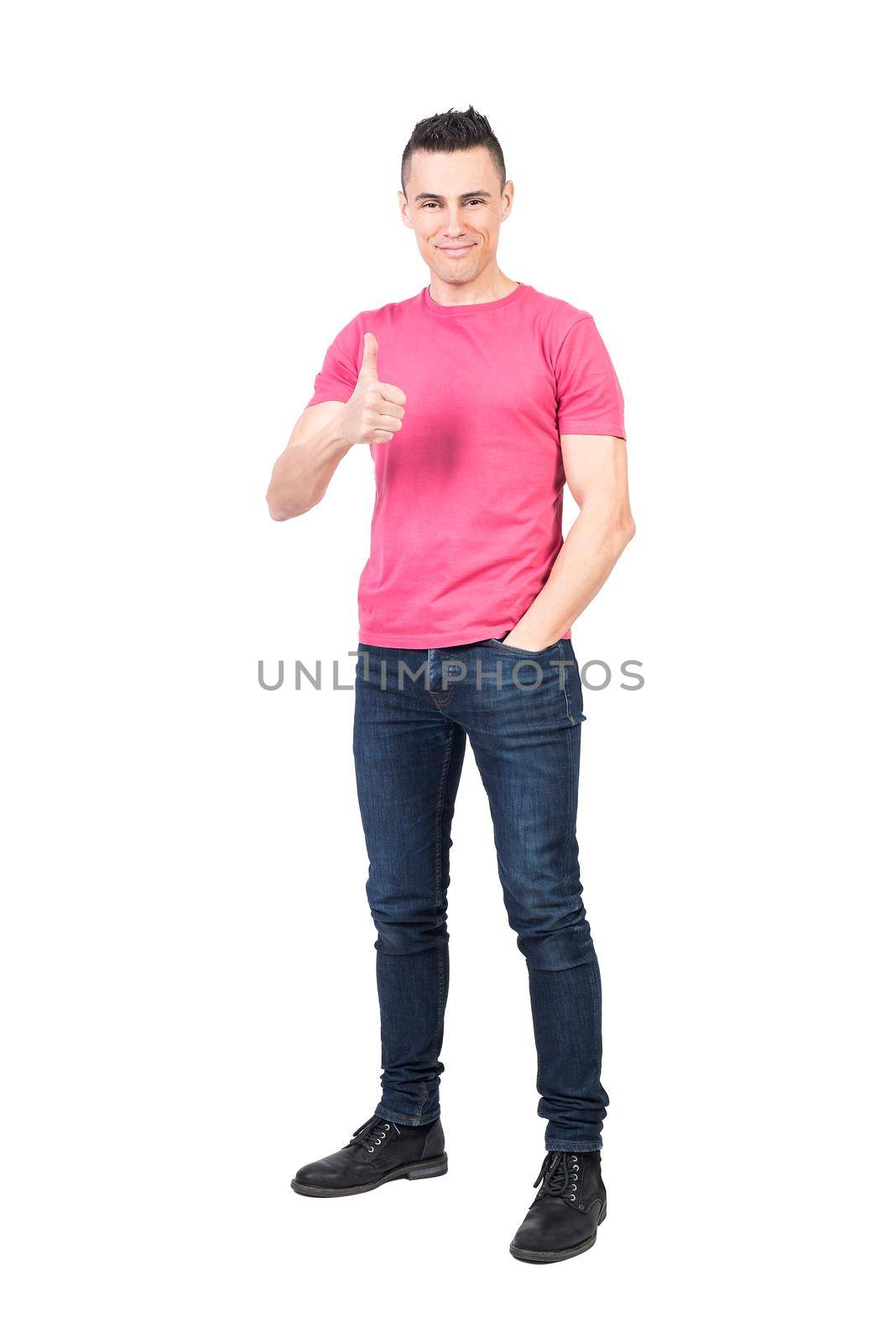 Full body of satisfied young male model with dark hair smiling and showing thumb up while standing against white background with hand in pocket and looking at camera