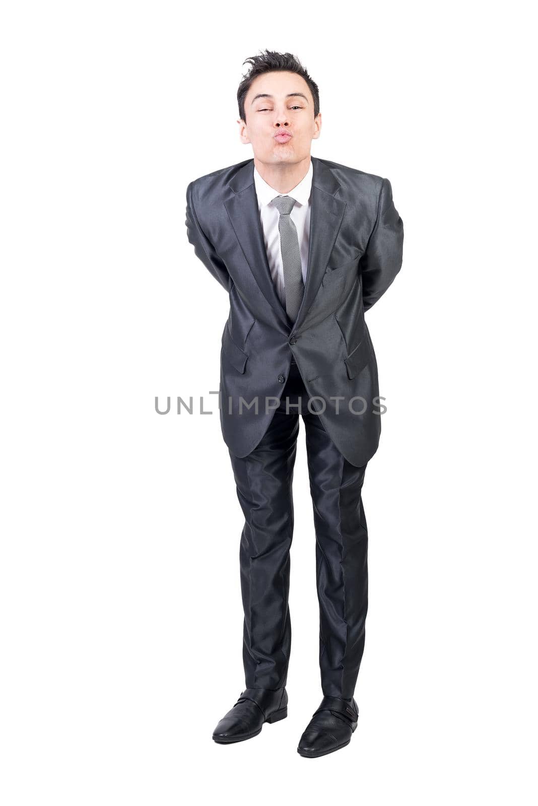 Full body of positive young guy with dark hair in elegant suit pouting lips and looking at camera while leaning forward with hands behind back against white background
