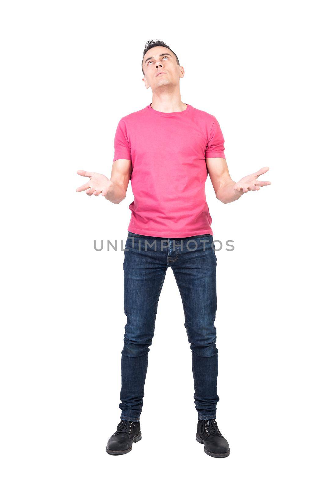 Full body regretful male in pink t shirt and jeans making helpless gesture and looking up while asking god for help in difficult situation isolated on white background