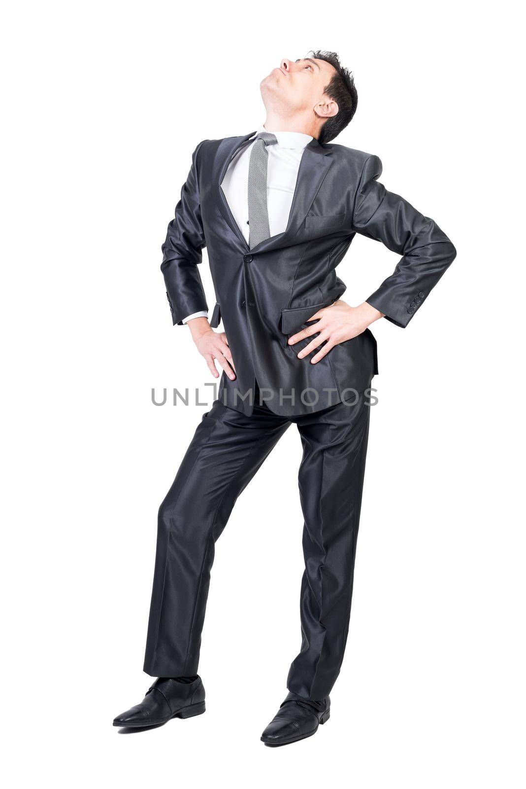 Full body of young arrogant male manager in classy suit and tie with dark hair standing against white background with hands on waist and looking up with confidence