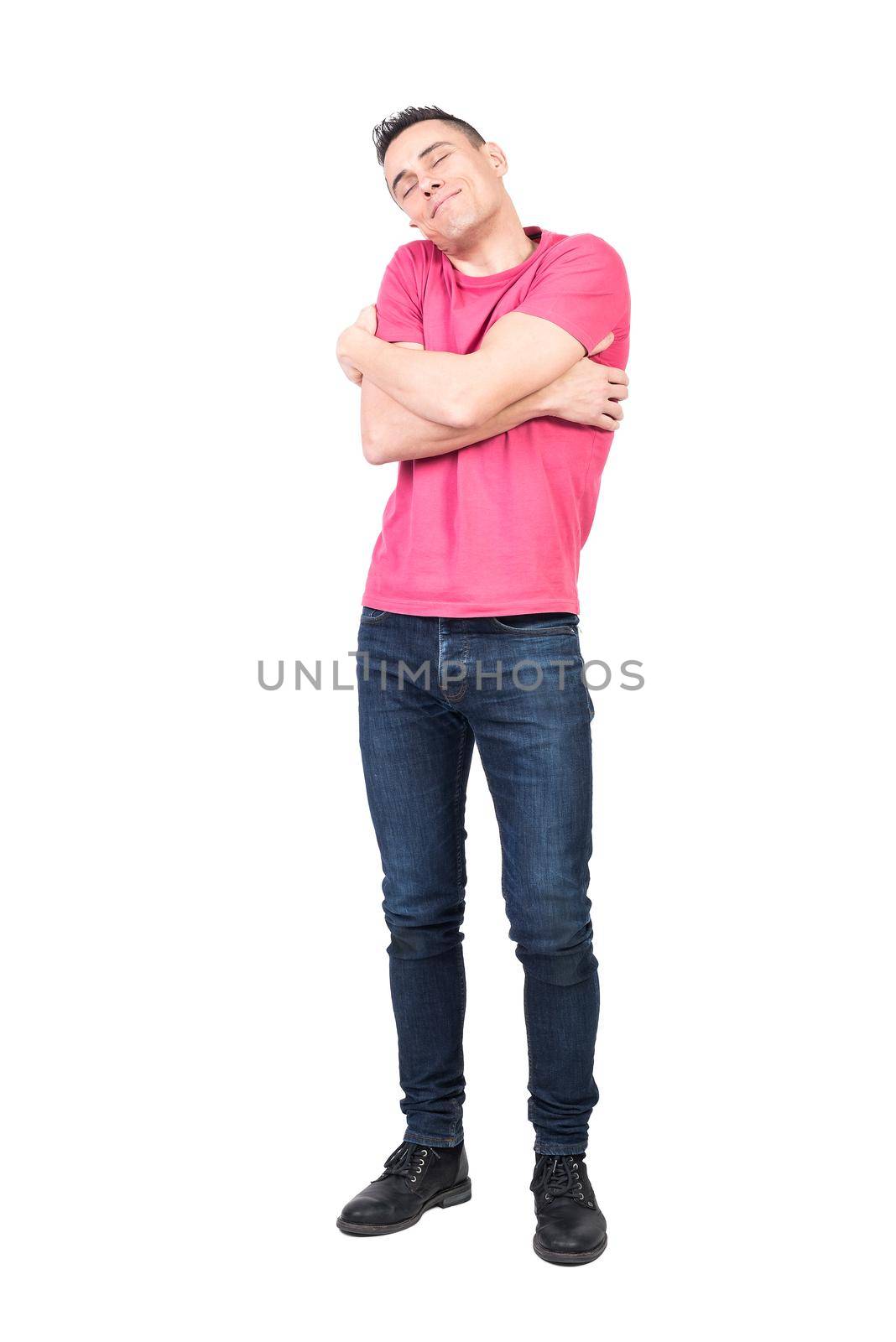 Full length of positive male in jeans and pink t shirt embracing shoulders and smiling with closed eyes while feeling love against while background