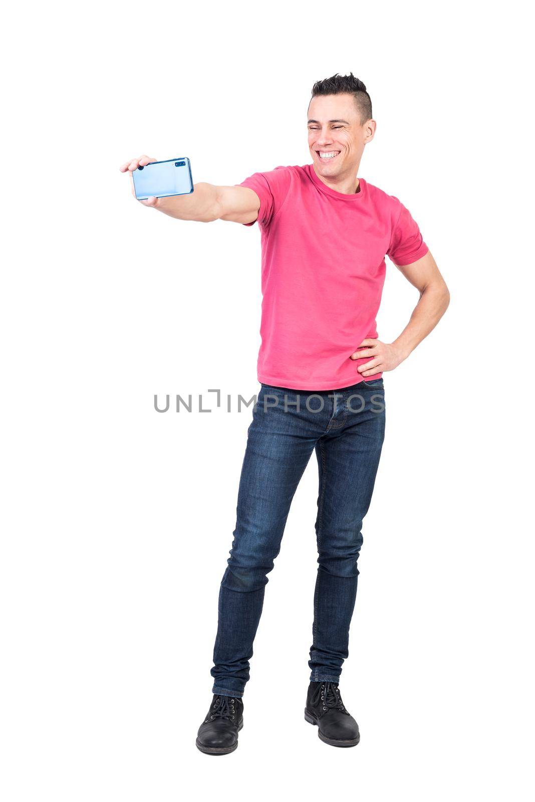 Full length of self assured young happy male with trendy haircut in casual outfit standing against white background with hand on waist and taking selfie on smartphone