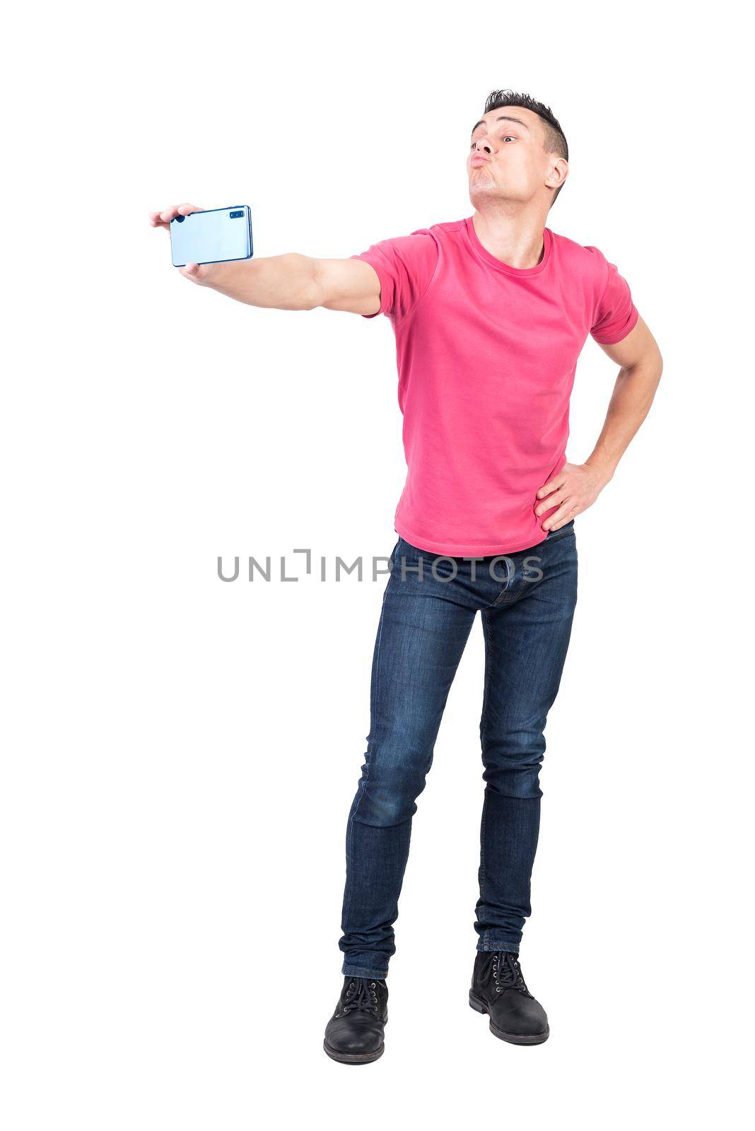 Full length of funny young guy with dark hair in t shirt and jeans pouting lips and taking selfie on mobile phone standing in studio with hand on waist