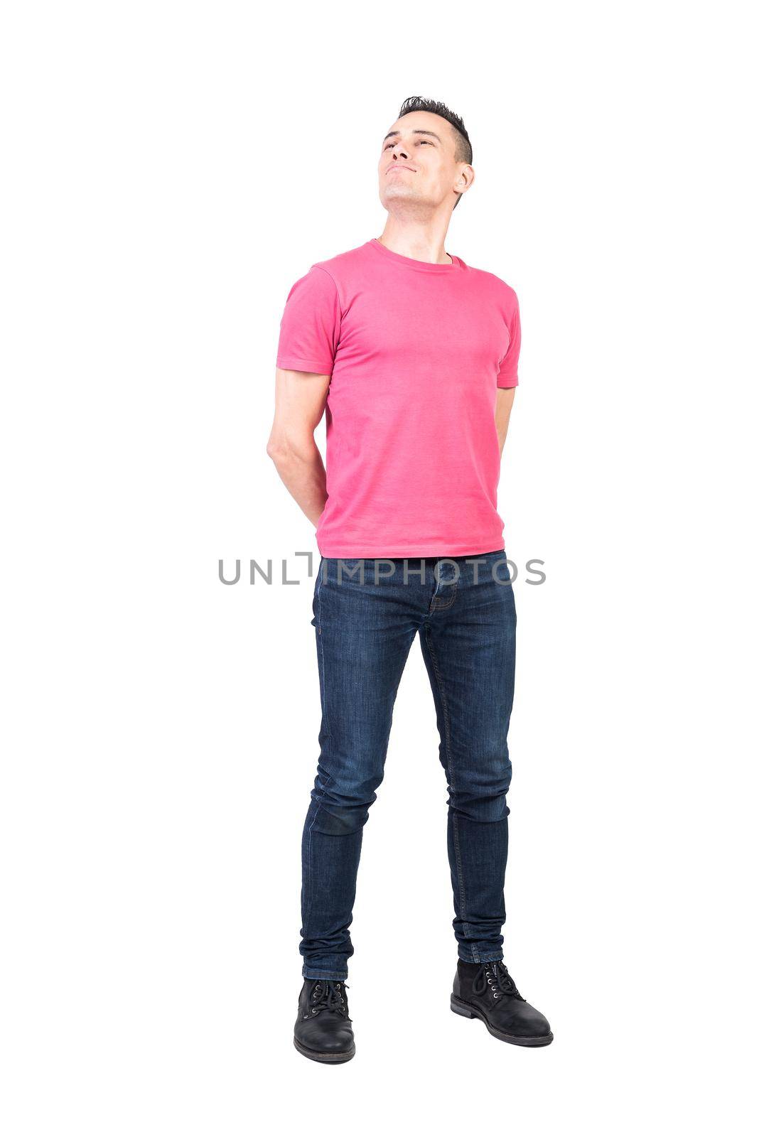 Full length of self assured young man in casual clothes standing in white studio with hands behind back and looking away proudly