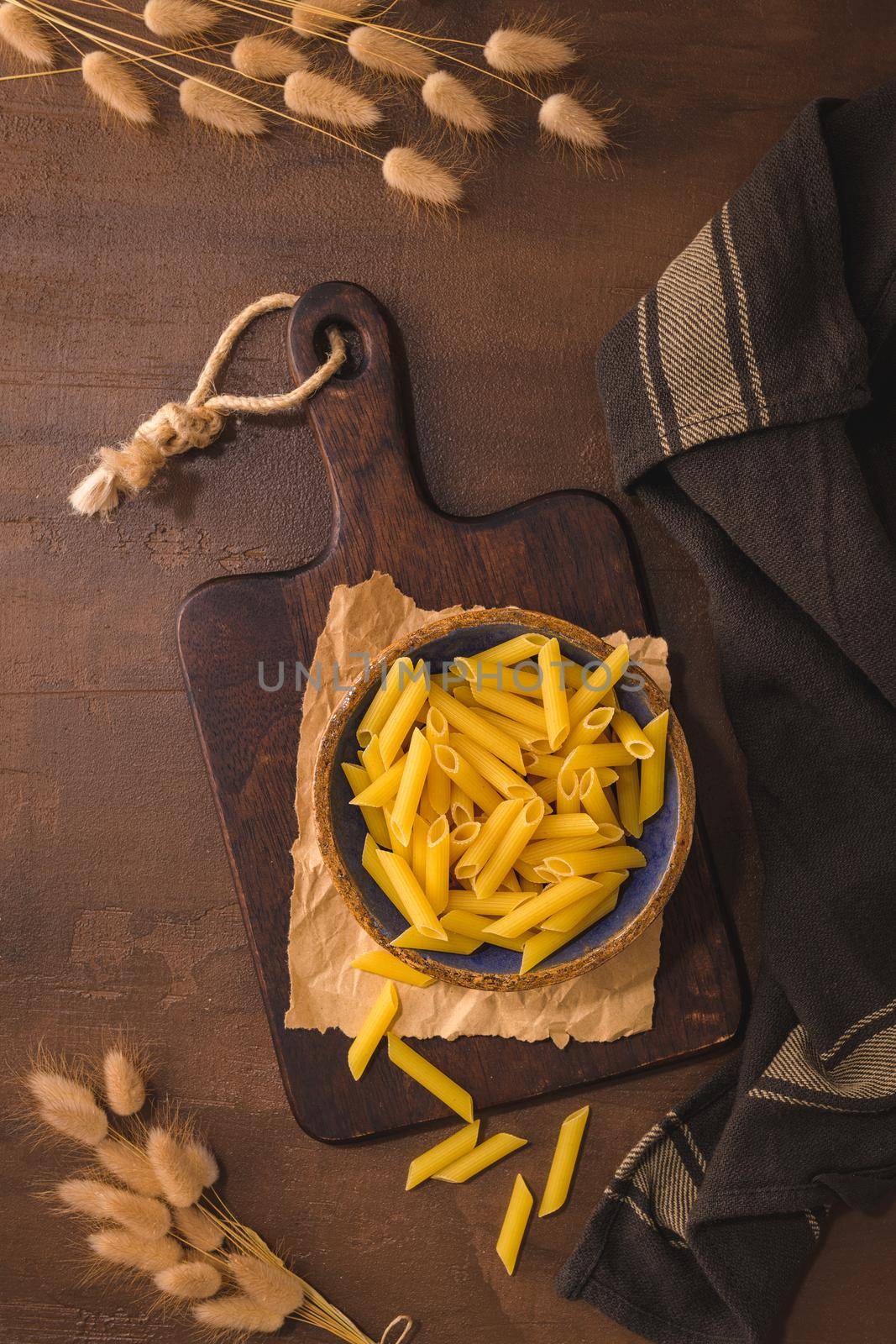Penne pasta in wooden cutting board on rustic kitchen countertop.