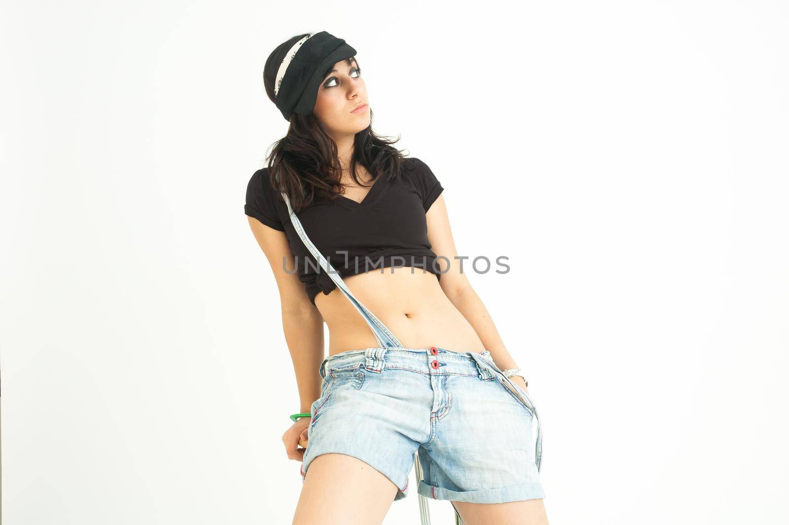 Modern pretty brunette girl with shirt, short jeans and cap sitting on a high chair looking sideways, on white background, mid shot