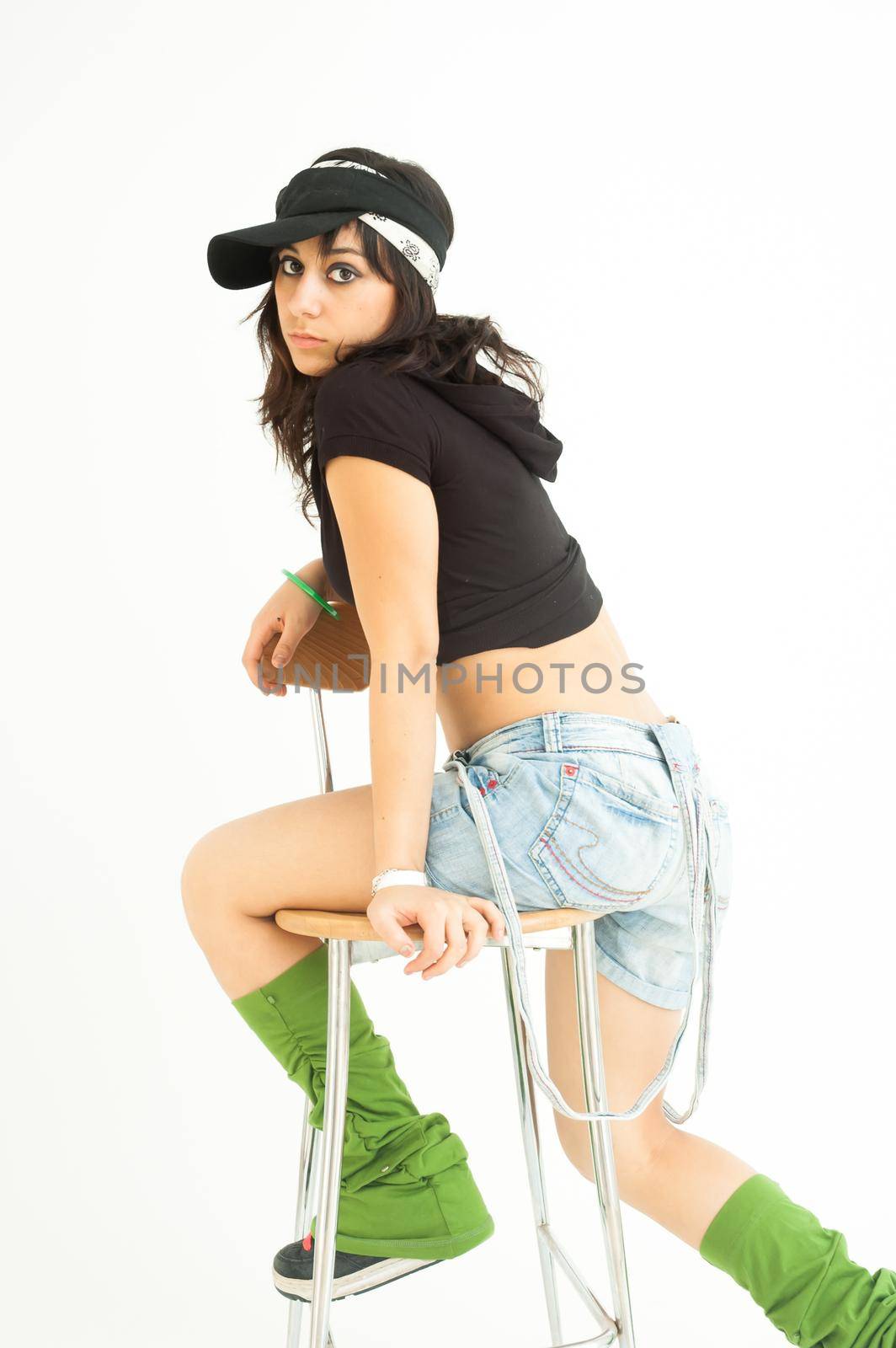 Modern pretty brunette girl with shirt, short jeans and cap sitting on a high chair looking at camera, on white background, long shot