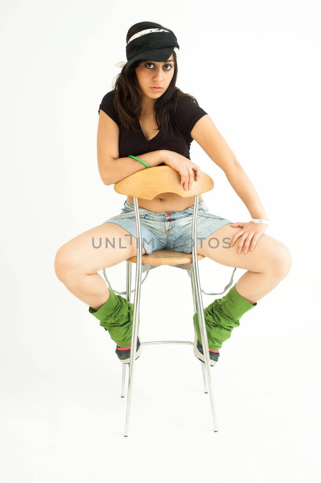 Modern pretty brunette girl with shirt, short jeans and cap sitting on a high chair staring at camera, on white background, long shot