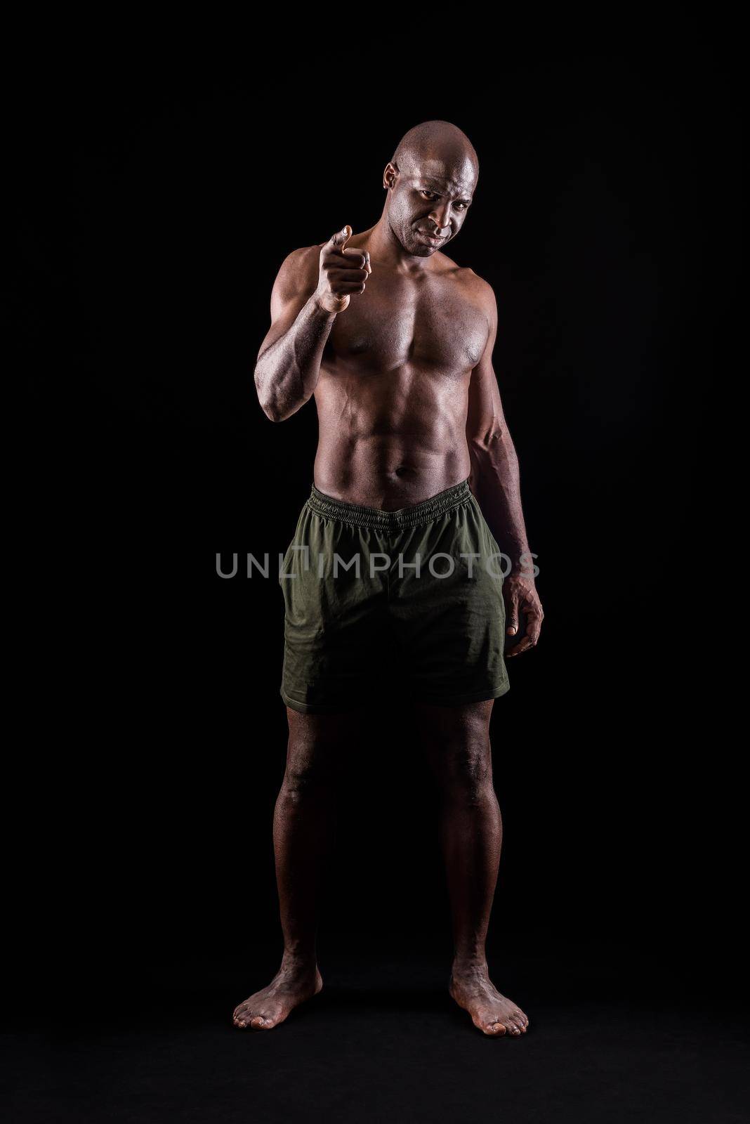 African american muscular man standing and pointing at camera with a confidence expression. Bodybuilder with bare torso and wearing shorts on a black background.