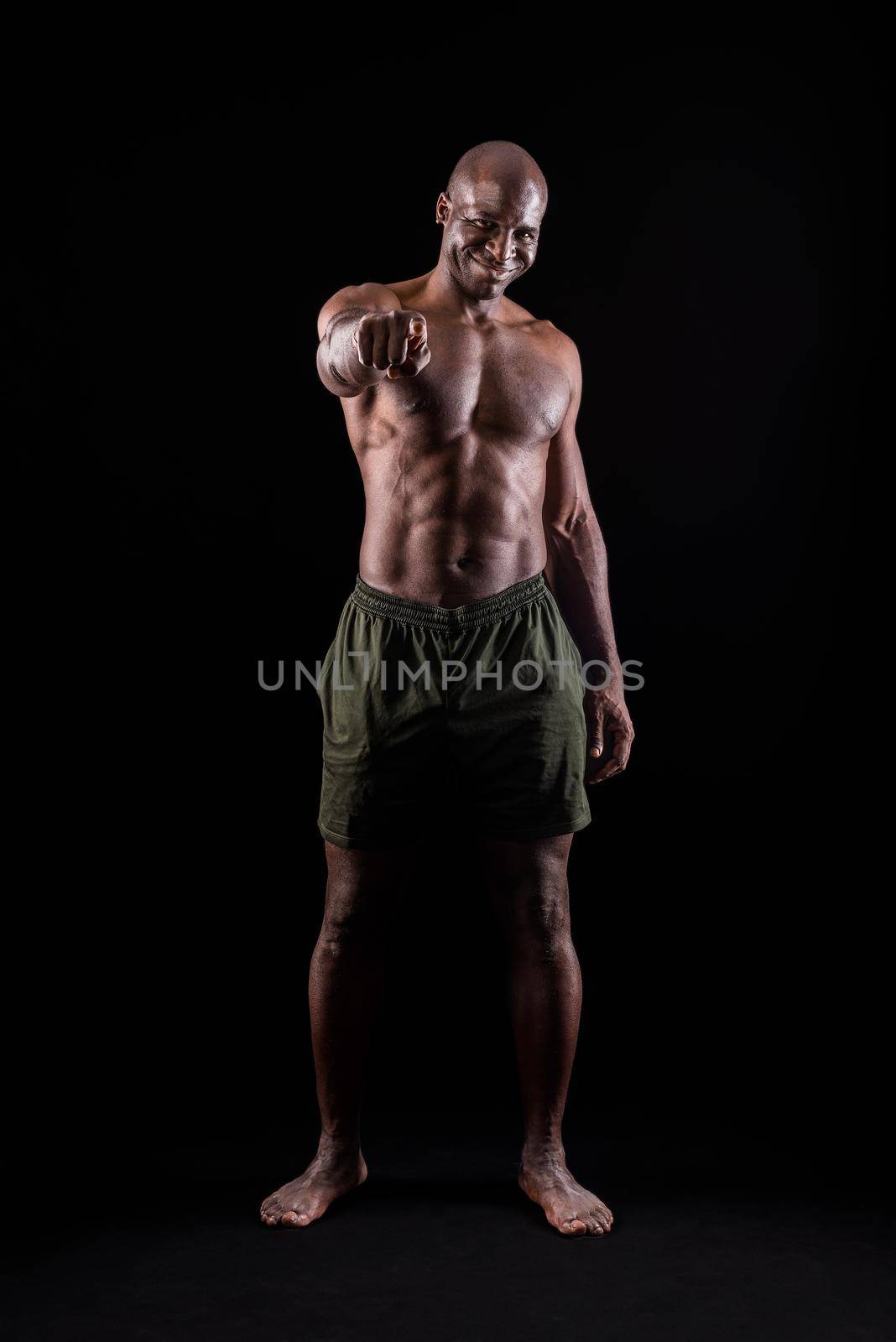 African american muscular man smiling standing and pointing at camera on a black background. Bodybuilder with bare torso wearing shorts in studio.