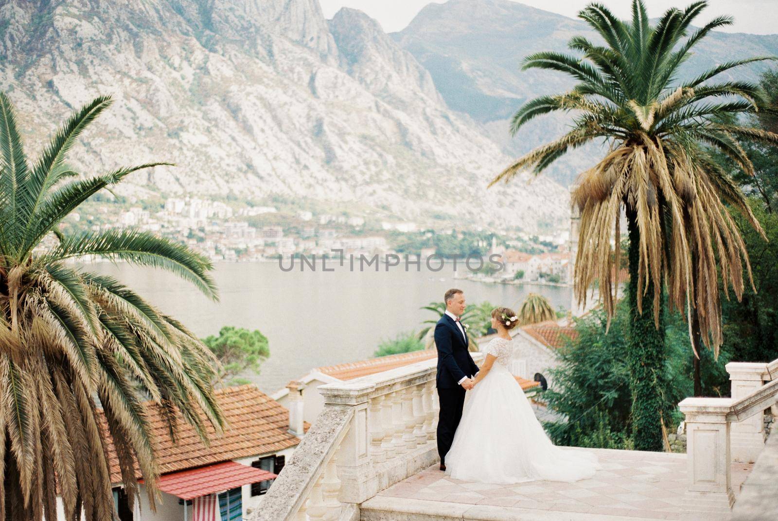 Bride and groom stand holding hands on the observation deck overlooking the sea and mountains. High quality photo