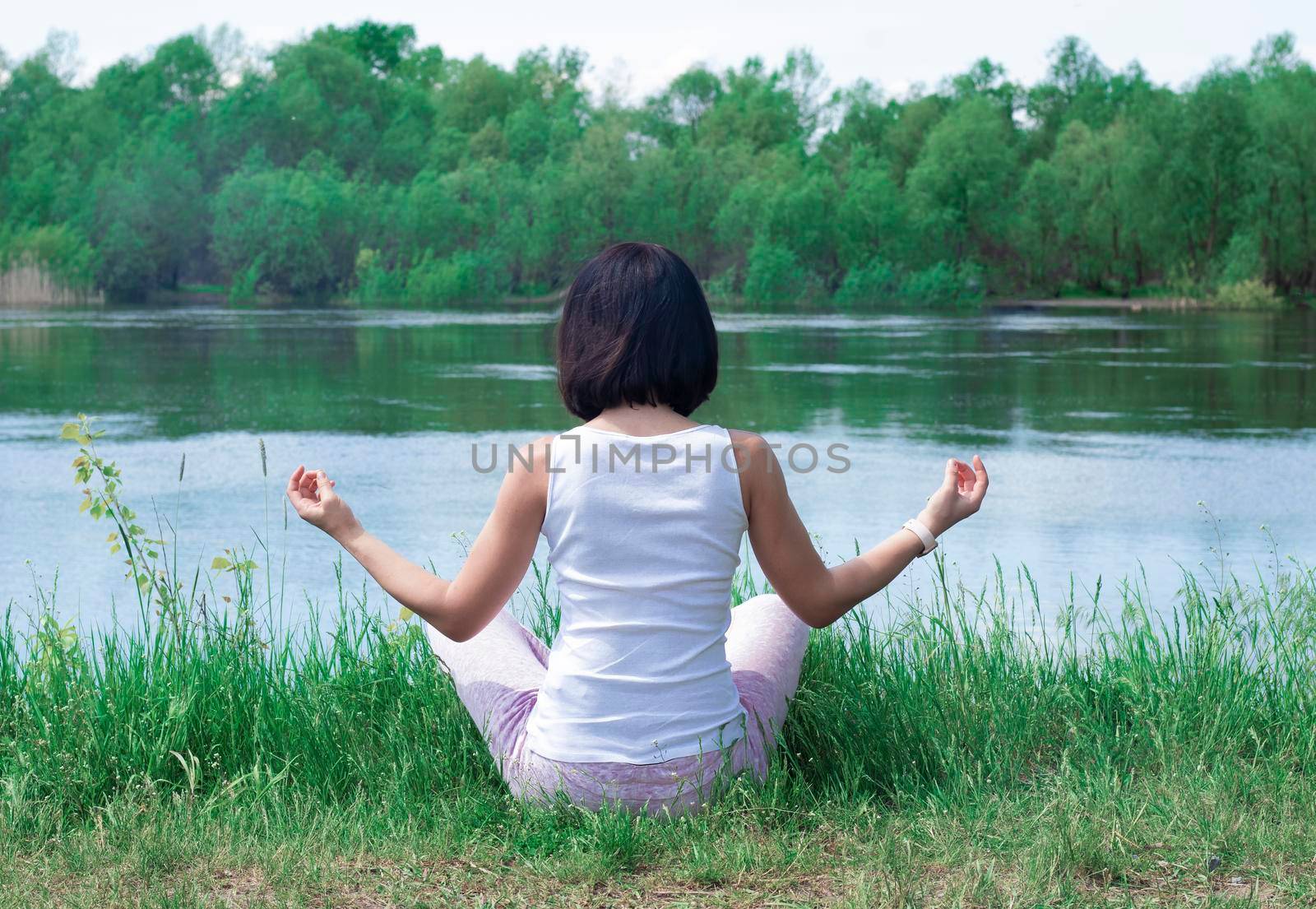 a girl sits in the grass by the river with her back in a meditate pose before. High quality photo. Yoga Concept. Hand Woman Practicing Lotus Pose. High quality photo