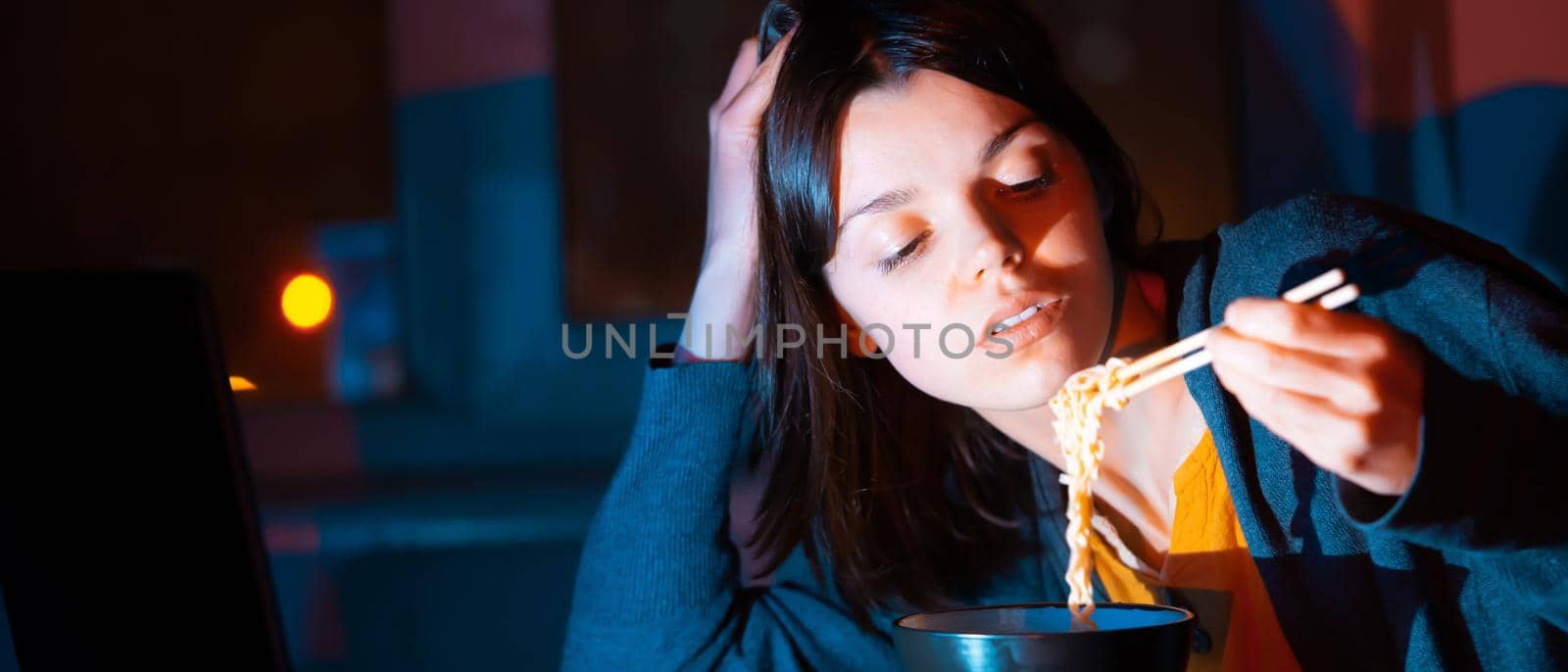 Young girl works at night at home with laptop and eats noodles, asian food with chopsticks. A woman watches a show, a movie and has dinner with ordered food from a cafe.