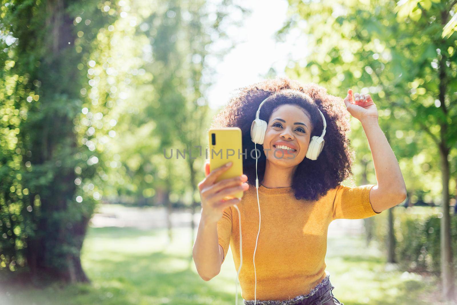 Pretty afro woman listens to music with headphones dancing in a garden. Selective focus.