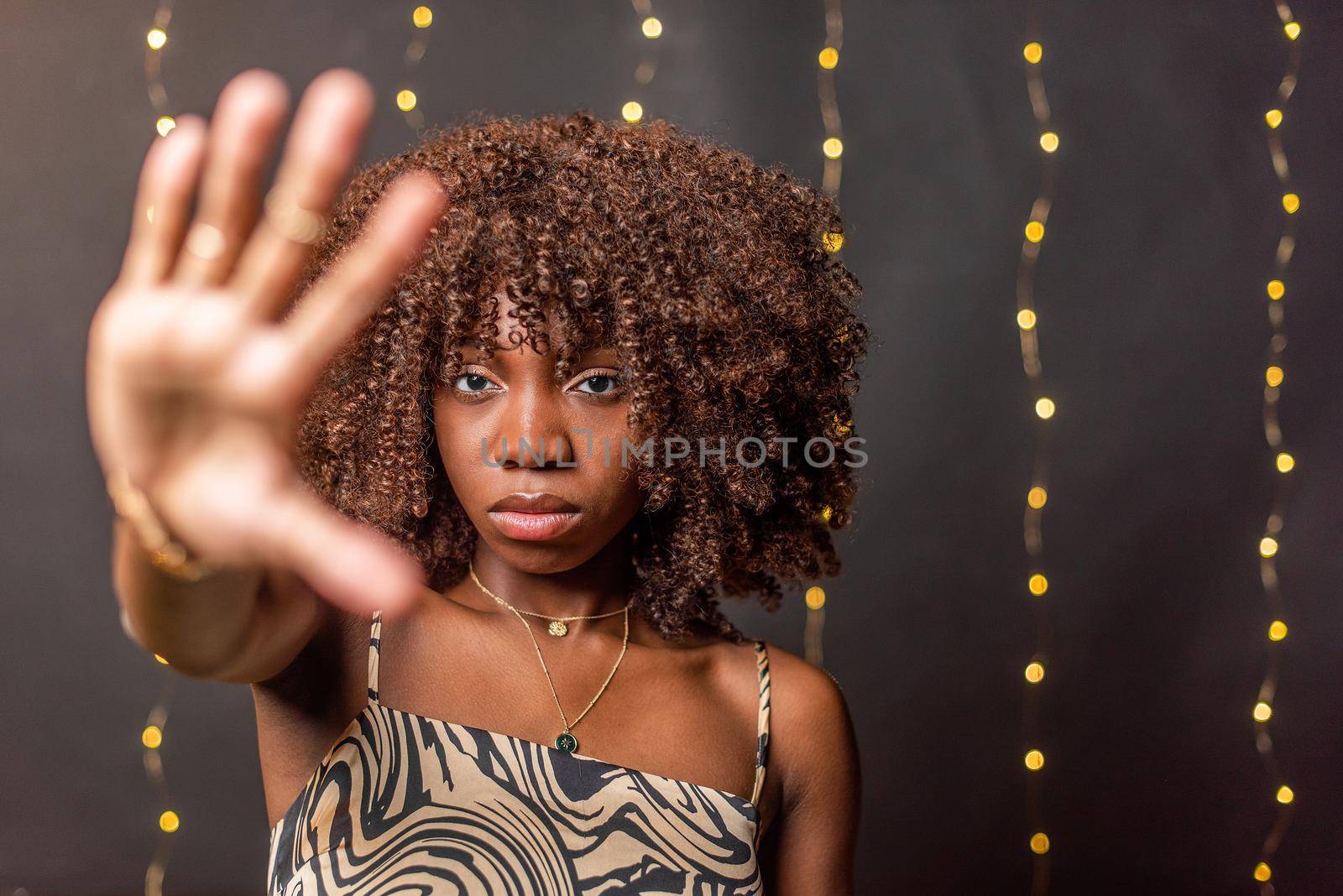 Unsmiling African American young woman with curly hair looking at camera showing palm on a blurred light background.