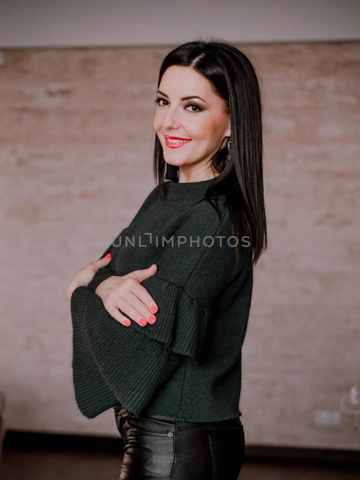 Portrait of adult beautiful mature woman standing on brown wall background. Lady in warm green sweater and leather pants with charming makeup and black hair smiling to camera.