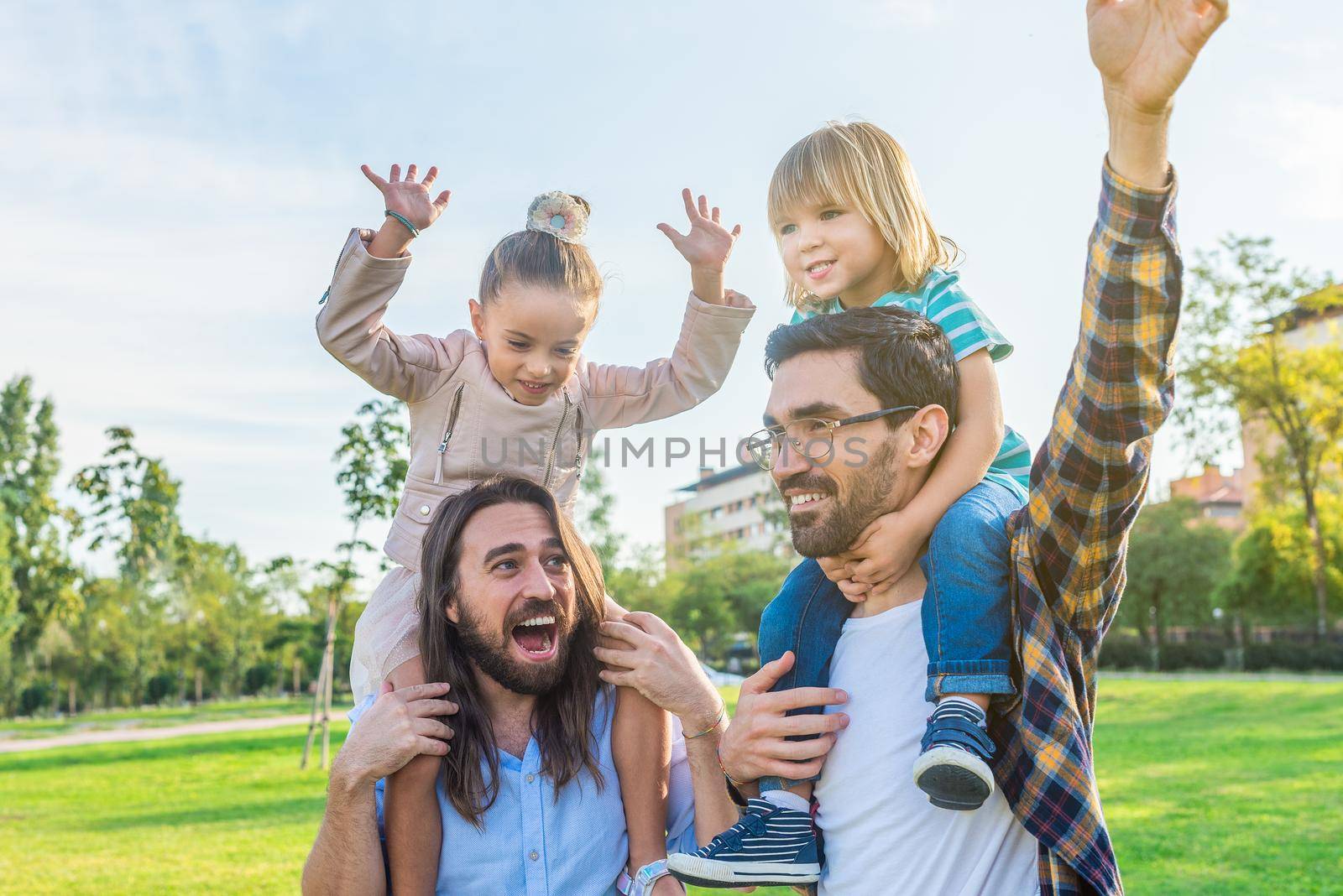 Two joyful caucasian dads holding their young children on their shoulders in the park in a sunny day.