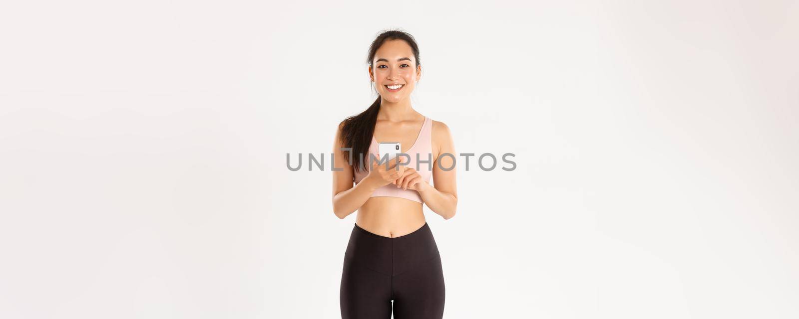 Sport, technology and active lifestyle concept. Smiling satisfied asian fitness girl, sportswoman holding smartphone, using running tracker app, checking heartrate during workout, white background by Benzoix