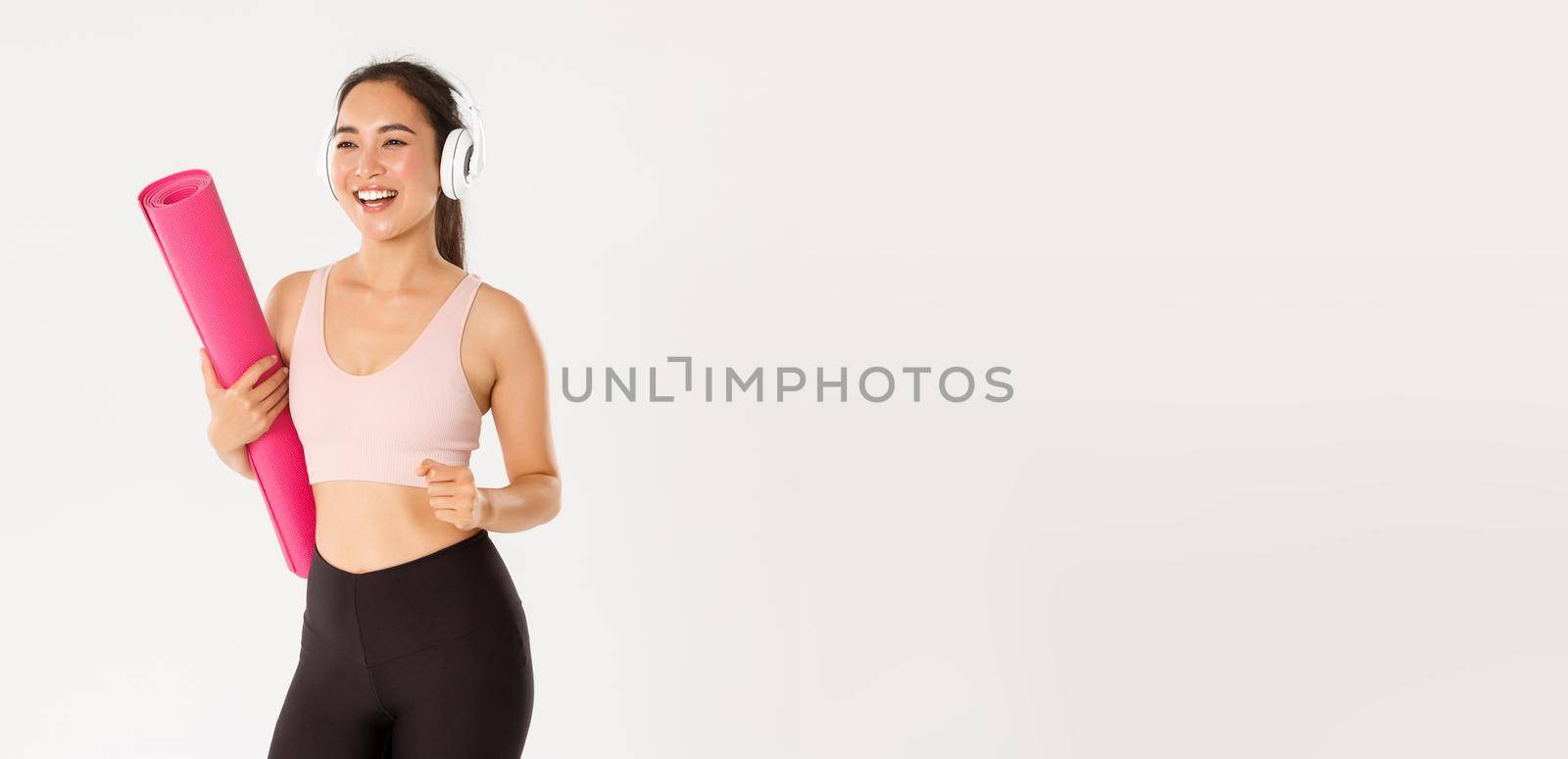 Sport, wellbeing and active lifestyle concept. Cheerful smiling asian fitness girl in headphones and sportswear going to gym with rubbermat, hurry up for yoga classes excited, white background.