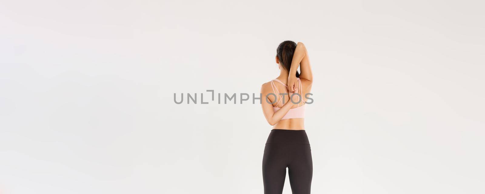 Full length rear view of active and slim brunette asian fitness girl, female athlete warm-up before yoga classes, lock hands behind back, sportswoman doing stretching exercises, white background.