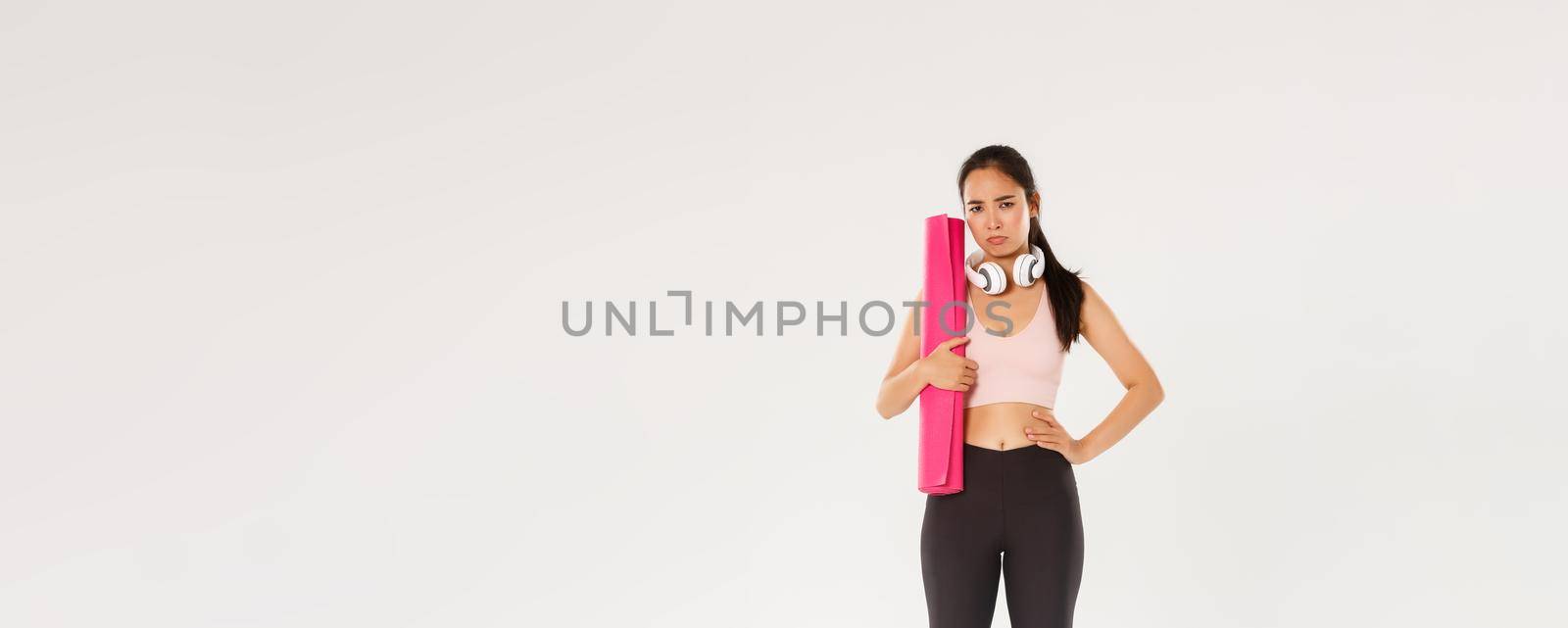 Full length of gloomy and upset cute brunette asian fitness girl in sportswear, sulking while holding rubber mat for yoga classes, looking disappointed as gym closed during covid-19 quarantine by Benzoix