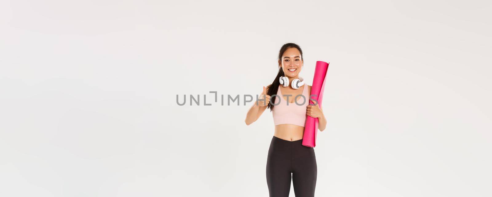 Full length of cheerful satisfied, slim asian girl with fitness rubber mat and headphones, female athlete showing thumbs-up, recommend gym or yoga classes, looking pleased after good workout.