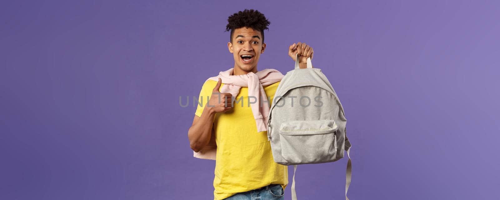 Portrait of cheerful young handsome male student, man recommending backpack, holding rucksack and show thumbs-up, bought new equipment for university semester, purple background.