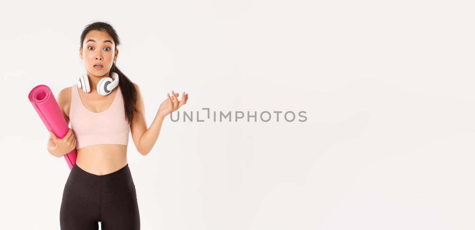 Sport, wellbeing and active lifestyle concept. Confused and shocked asian fitness girl, cant understand why gym closed, holding rubber mat and looking surprised at camera, white background.