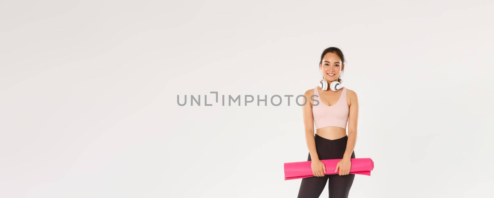 Full length of smiling slim asian girl with rubber mat for fitness exercises, headphones, looking at camera while waiting for training coach starting workout session in gym, white background.