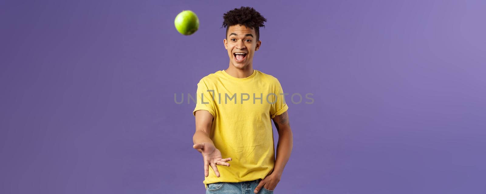 Holidays, vitamins and vacation concept. Portrait of handsome upbeat young male student asking friend something eat, catching apple and smiling happy, standing purple background by Benzoix