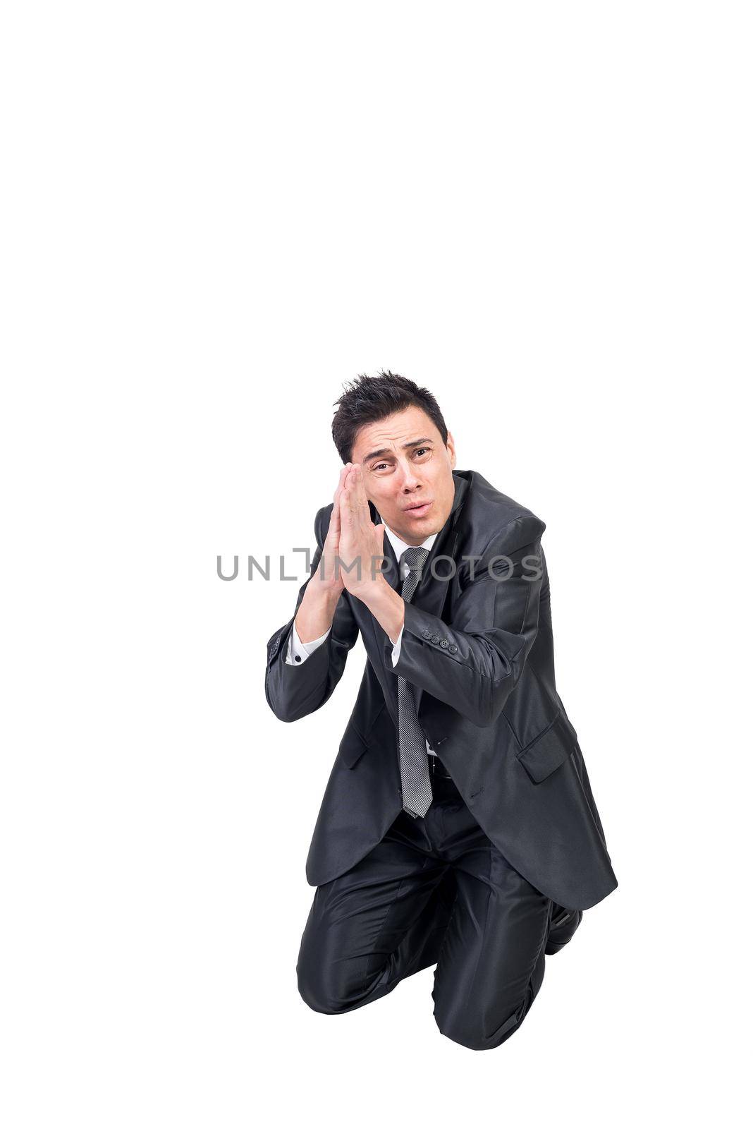 Full body of unhappy male in formal suit knelling on floor and supplicating with prayer hands while looking at camera in studio against white background