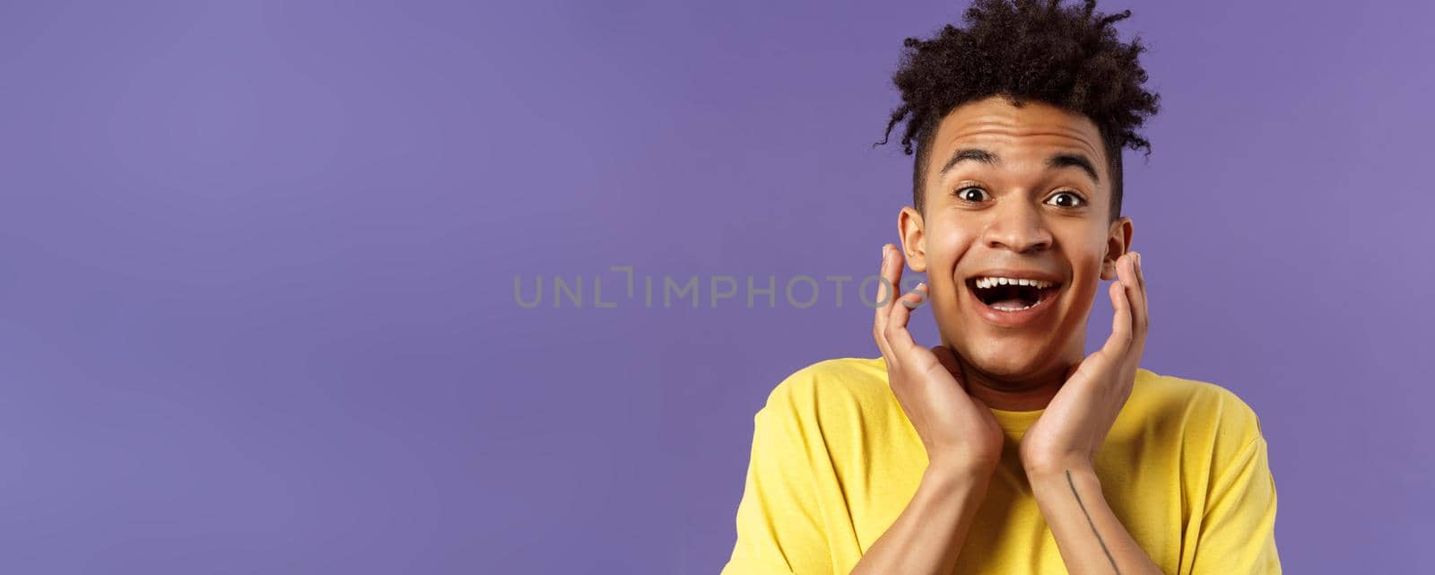 Close-up portrait of extremely happy, enthusiastic young man hear fantastic news, looking surprised and excited, touching face in joy, smiling upbeat look camera astonished, purple background.