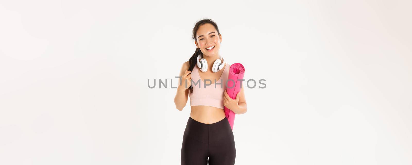 Sport, wellbeing and active lifestyle concept. Smiling happy asian fitness girl in headphones and sportswear, carry rubber mat for workout or yoga, laughing carefree over white background by Benzoix