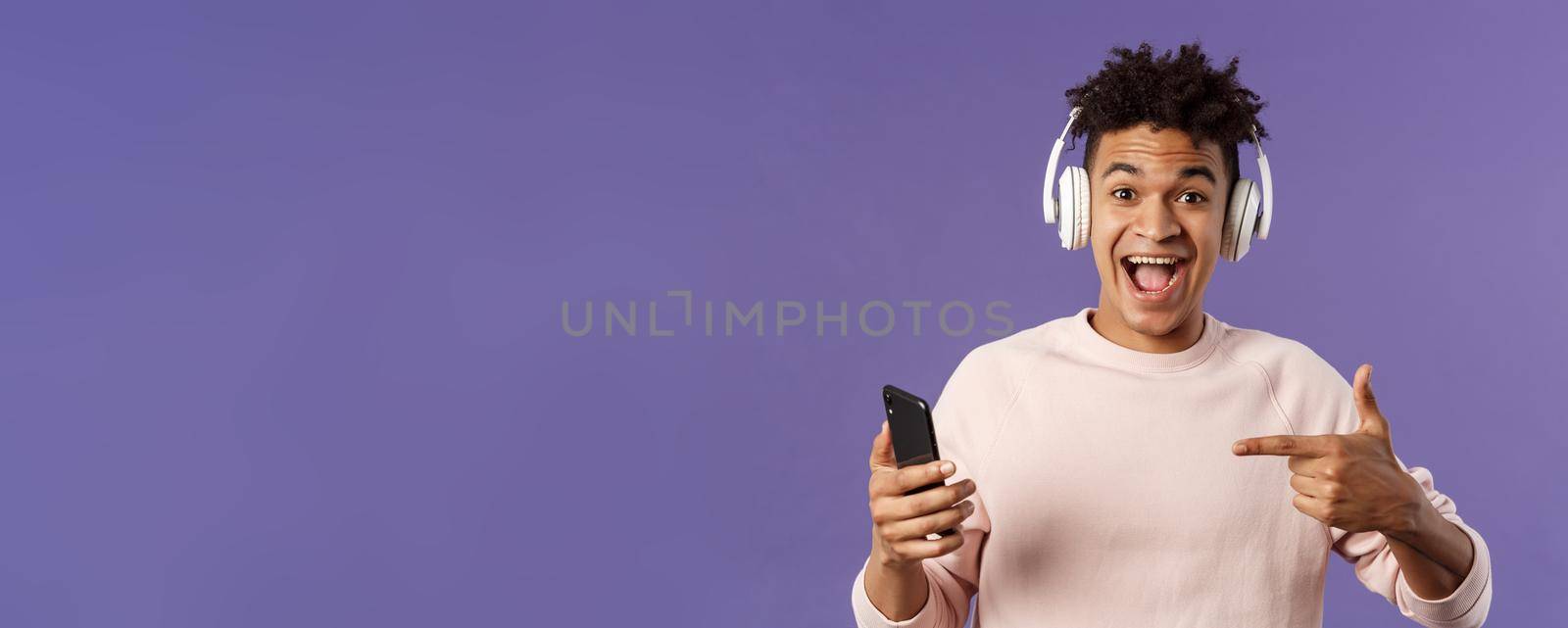 Technology and lifestyle concept. Portrait of happy, cheerful young man recommend awesome podcast or online music platform, buying subscribtion listen songs anytime, wear headphones, point at phone by Benzoix