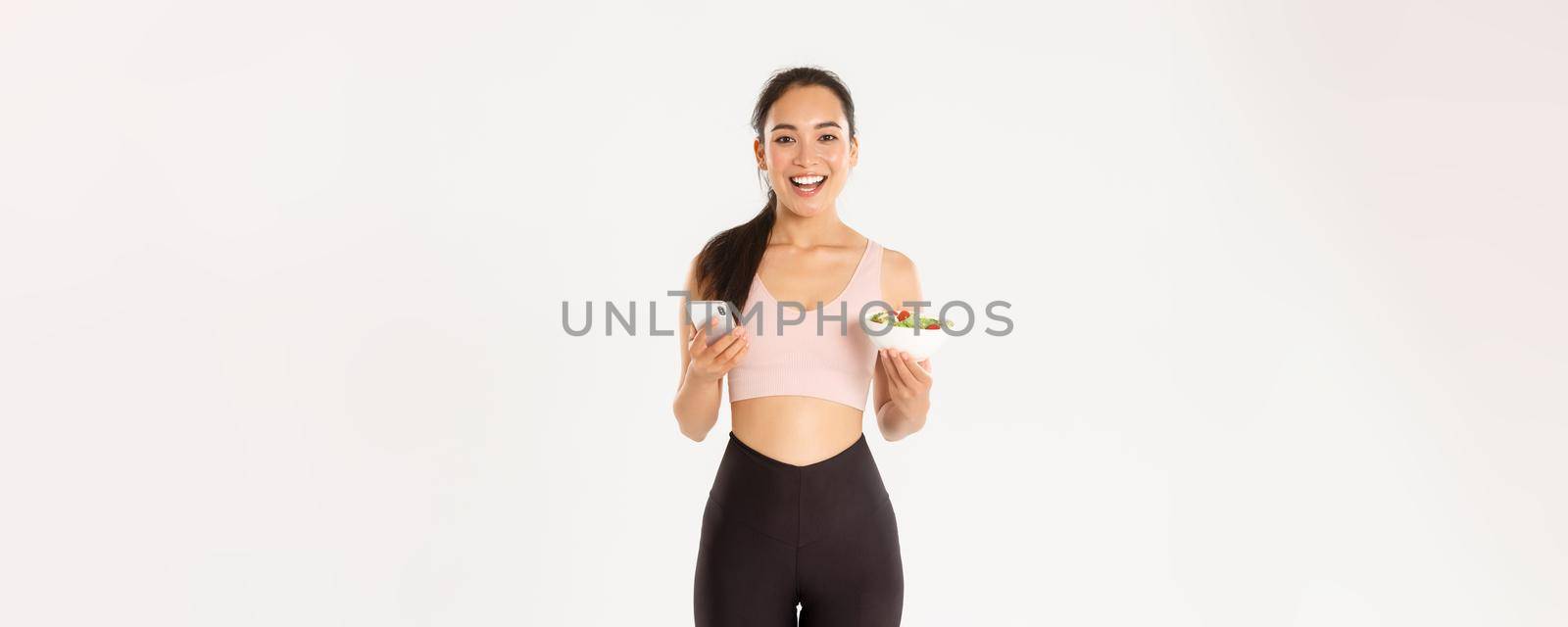 Sport, wellbeing and active lifestyle concept. Smiling slim asian fitness girl in sportswear, holding salad and mobile phone, using eating reminder app, diet control application, checking calories by Benzoix