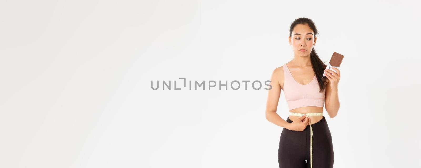 Sport, wellbeing and active lifestyle concept. Thoughtful and indecisive asian girl looking gloomy over body weight, measuring waist but looking at chocolate bar, pondering, white background.