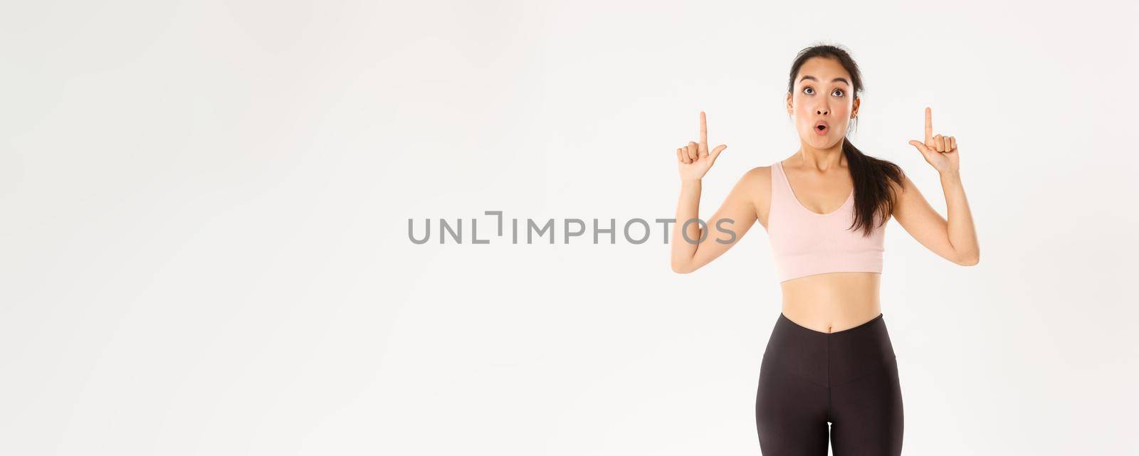 Sport, wellbeing and active lifestyle concept. Excited and happy asian fitness girl in sportswear, pointing fingers up and looking amazed with great discounts, read banner, white background.