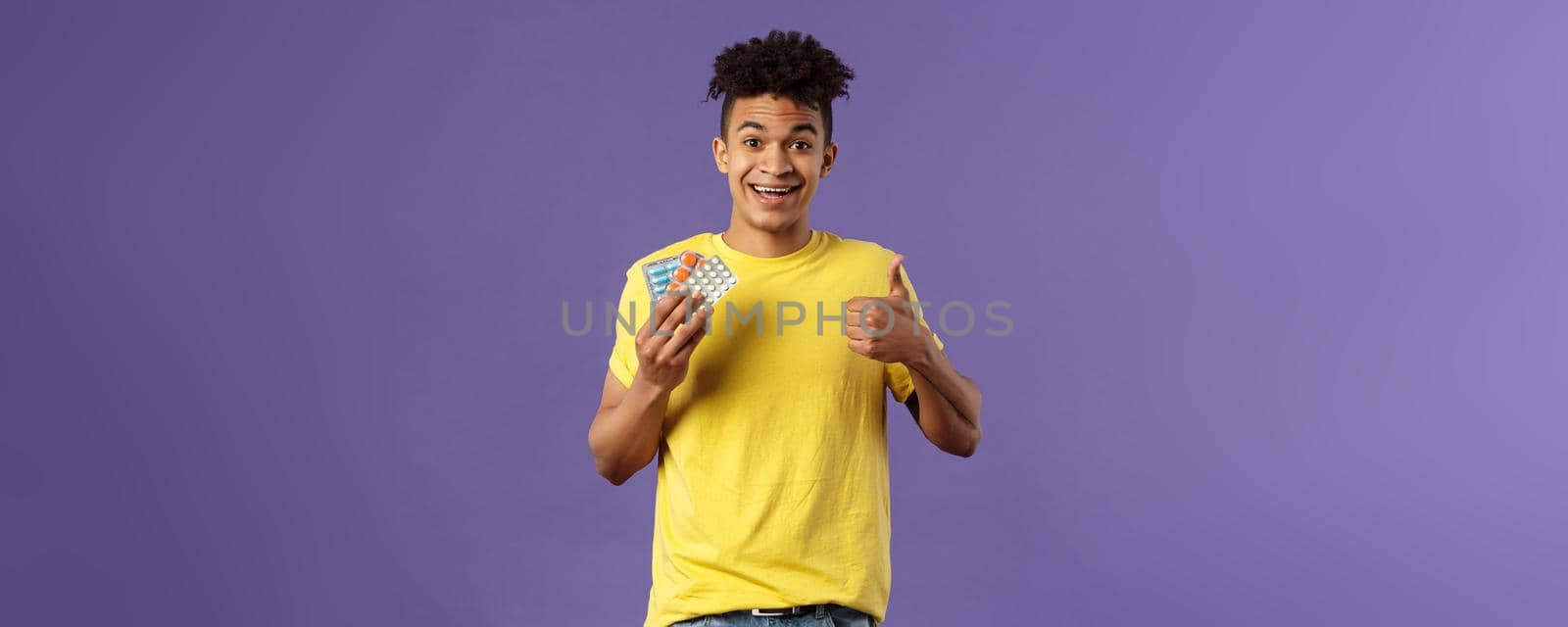 Health, influenza, covid-19 concept. Portrait of young healthy man got better, show thumbs-up holding drugs, taking pills to feel better after catching cold, being sick, purple background.