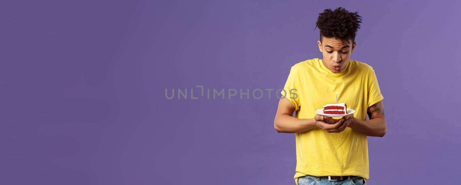 Portrait of cute excited young guy with dreads and tattoos, making wish on birthday, celebrating party, blowing candle on b-day cake, standing purple background dreaming. Holidays concept