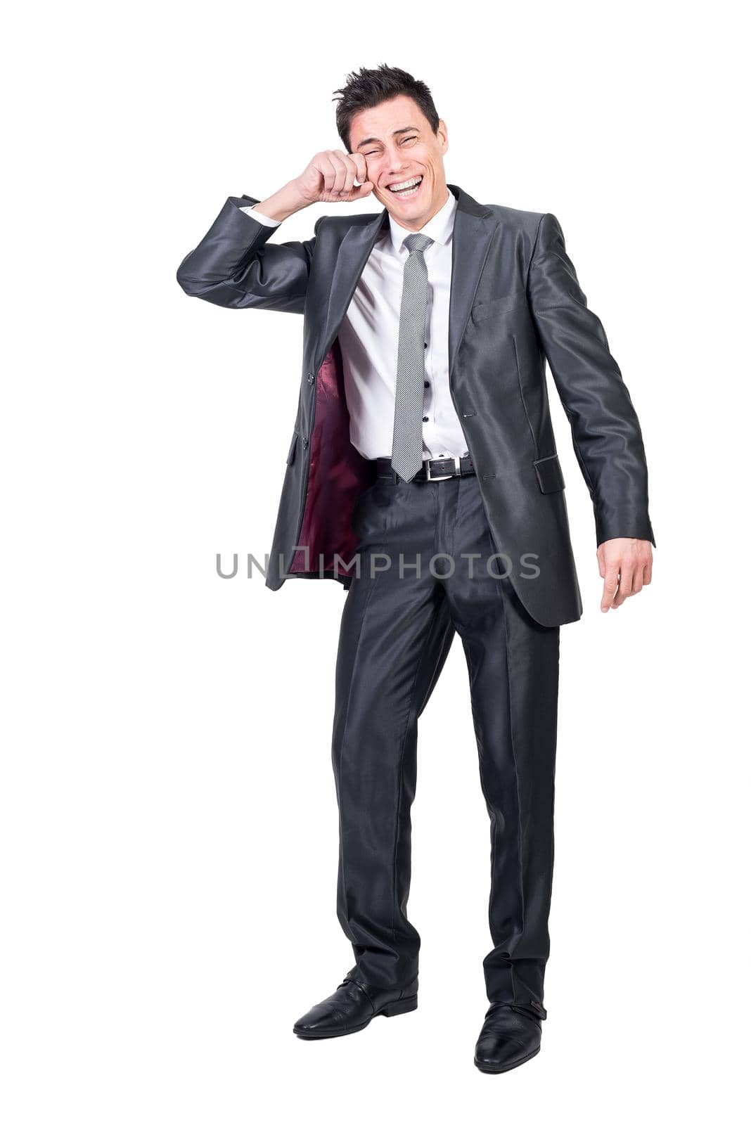 Full body of joyful man in formal suit laughing on funny joke and wiping tears from face in studio against white background
