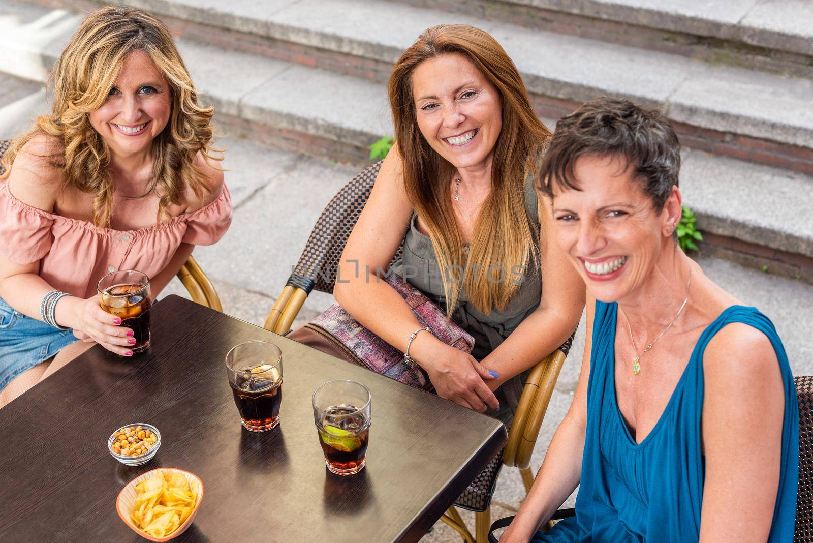 Three adult women in a cafe outside looking at camera laughing and having drinks. High angle view of beautiful mature women having a good time together.