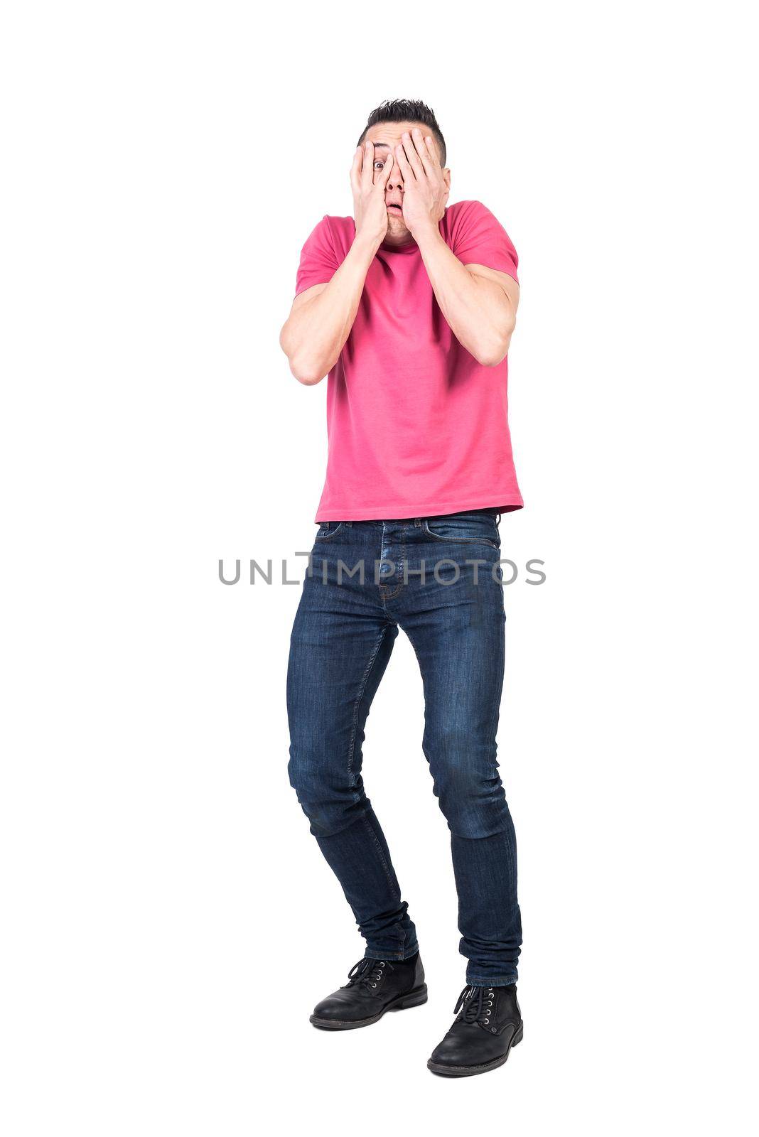 Full length scared man in casual clothes covering face with hands and peeking through fingers isolated on white background