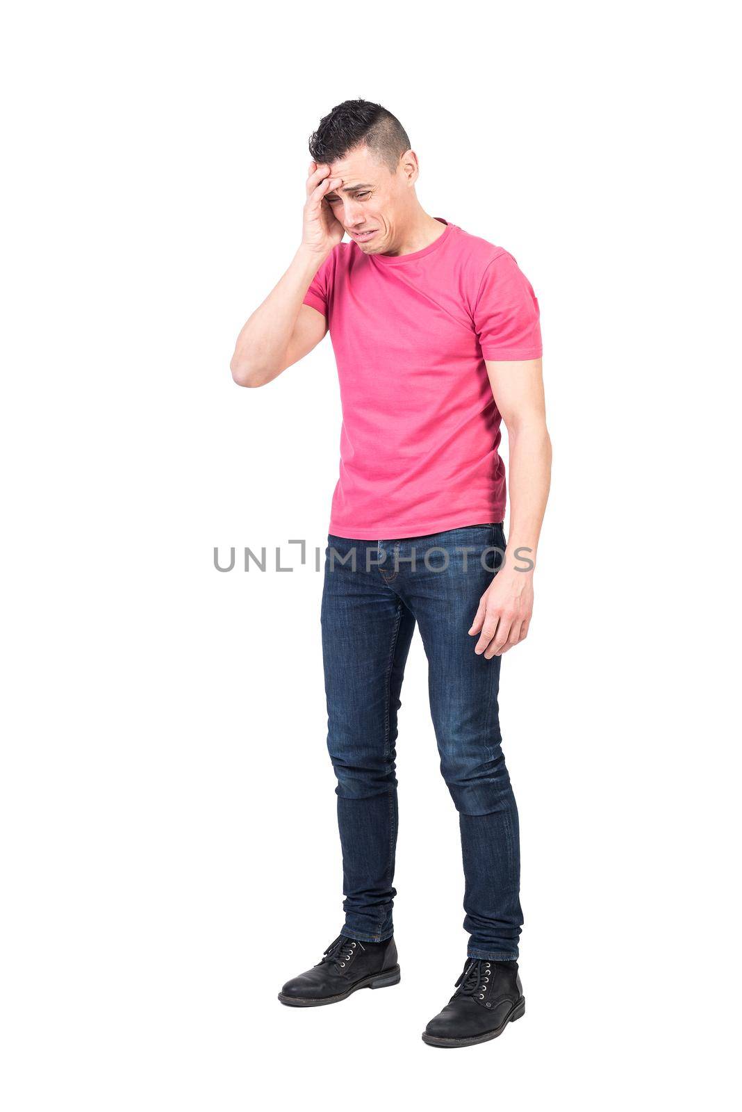 Full body of young upset male with dark hair in pink t shirt and jeans touching forehead and looking down with disappointment while crying against white background