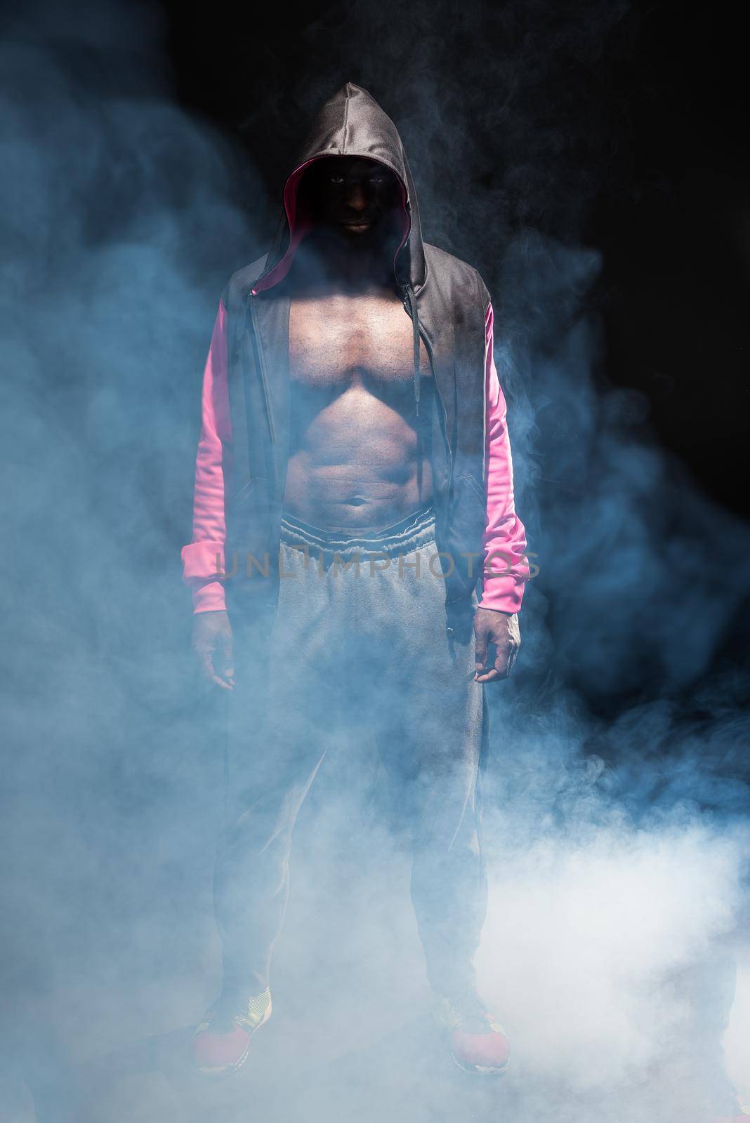 Muscular adult male in hooded jacket with bare torso with smoke around. African American man in sportswear standing in a studio with black background.