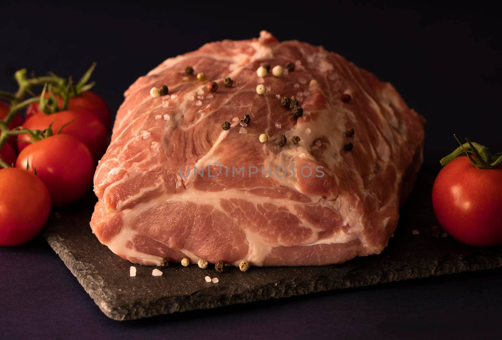 large raw piece of meat is seasoned with a mixture of peppers and salt. Lies on a stone board with fresh tomatoes. Pork marinated with spices. Fresh meat is ready to cook. High quality photo