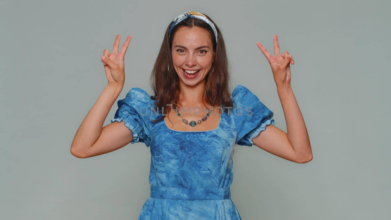 Lovely happy young woman showing victory sign, hoping for success and win, doing peace gesture, smiling with kind optimistic expression. Adult stylish girl isolated on gray studio background indoors