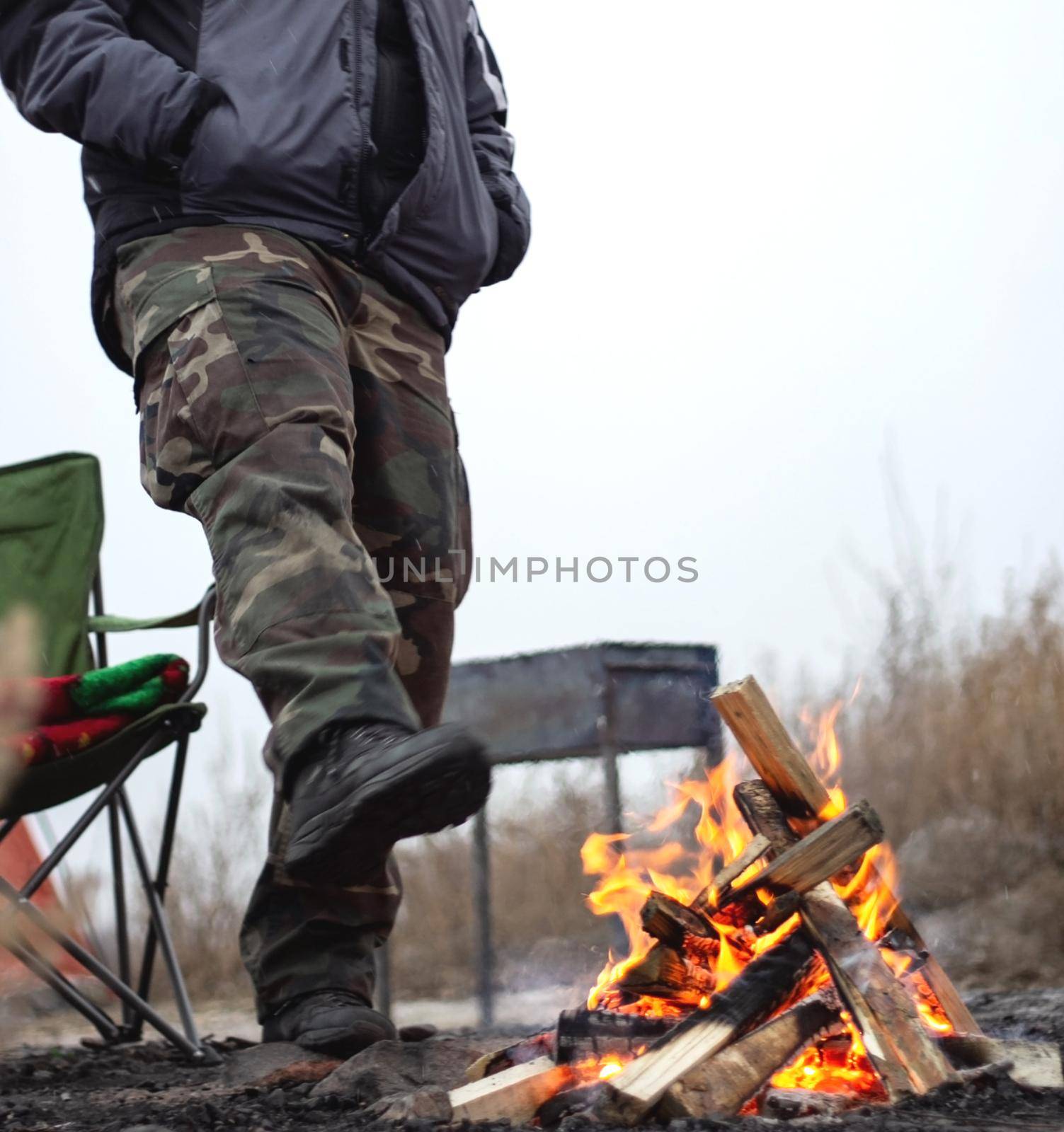 Faceless man in traveling clothes get warm stending by the fire in late autumn or winter with first snow, authentic extreme camping in cold cloudy overcast weather, vertical image with low angle